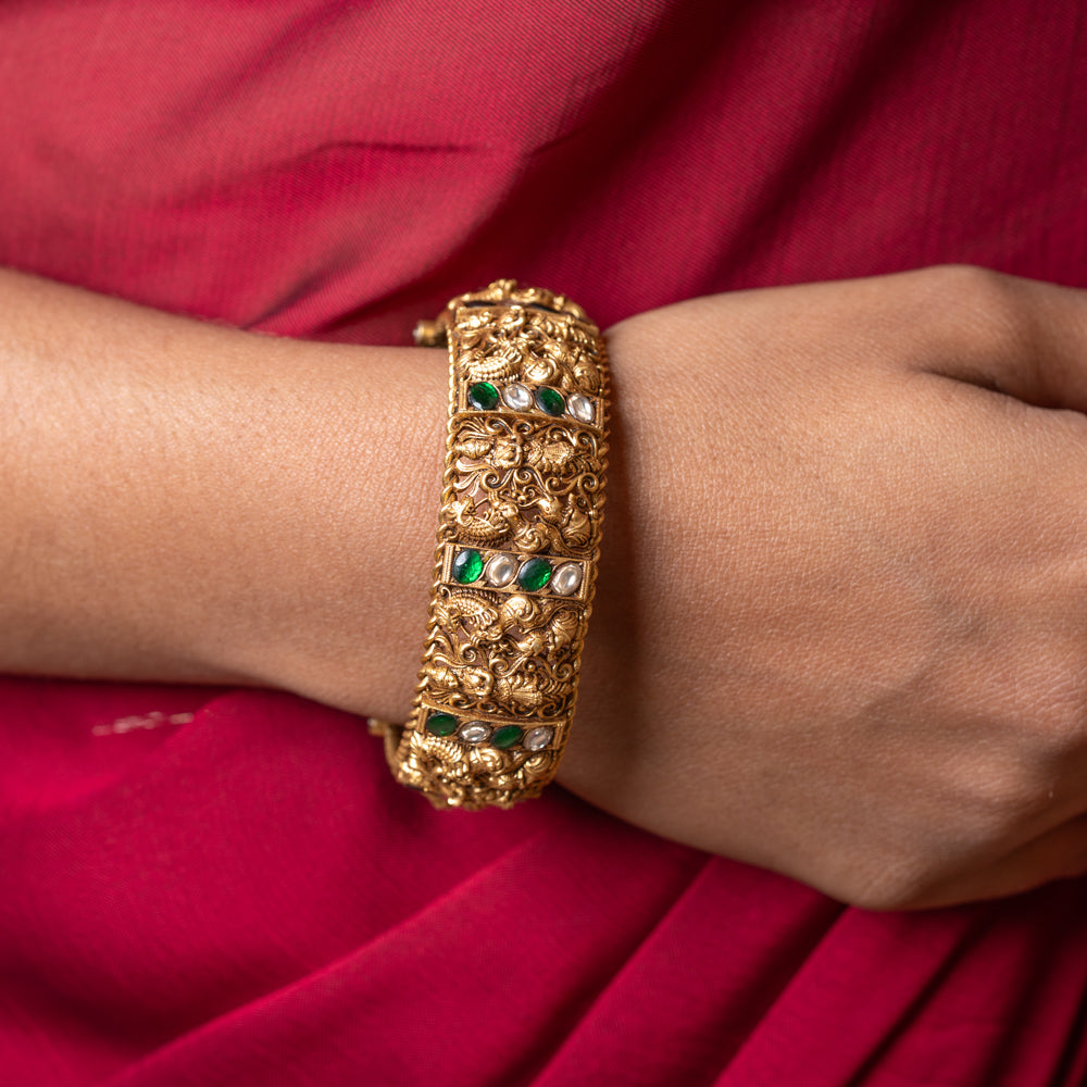 Gold bracelet with green and red stones on a wrist against a red fabric background